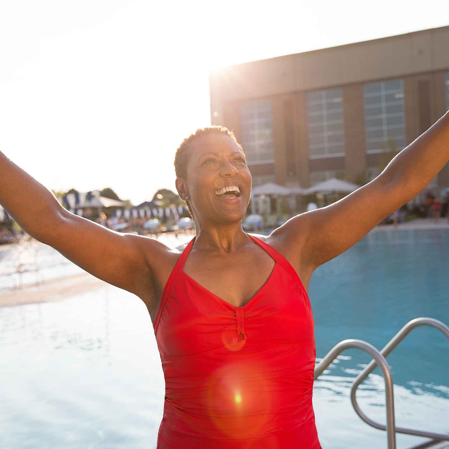 A smiling photo of a middle-aged woman from the chest up, her bare arms raised victoriously in a “V” formation. The woman, who is African American, is outside on one of Life Time’s pool decks.